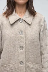 Trench de lana fina sin teñir, con diseño de textura combinada. Presenta mangas y espalda tejidas con patrón de trenzas, cuerpo liso, cuello camisero, cierre frontal con botones y bolsillos delanteros funcionales.
