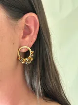 Aro de metal dorado de 2 cm de diámetro, con forma circular, decorado con cuentas de vidrio facetadas en tonos ámbar y marrón oscuro.