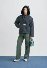 Campera con capucha Carhartt WIP modelo Mitch, color marrón oscuro, confeccionada en lona de algodón de peso medio con forro interno de corderito sintético. Cuenta con cierre frontal bidireccional, bolsillos frontales tipo parche y etiqueta cuadrada tejida con el logo de la marca.