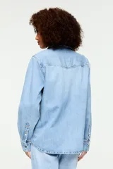 Camisa de denim liviano de color azul claro, con inspiración western, cuello clásico, cierre frontal con broches metálicos y bolsillos delanteros con solapa. Presenta un calce relajado y mangas largas con puños abotonados.