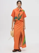 Falda larga pareo color anaranjado con estampado difuminado en tonos verdes y azules, tiro alto y tajo delantero.