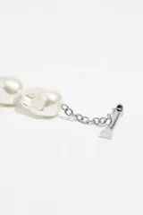 Collar de diseño mixto que combina una sección de perlas irregulares con una sección de cadena de eslabones dorados. Incluye un cierre tipo barra en T y dos dijes colgantes en forma de corazón, uno dorado con logo grabado y otro esmaltado en negro.