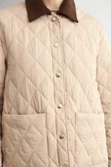 Campera capitoneada de diseño acolchado con patrón de rombos, color beige claro. Presenta cuello camisero de pana en color marrón oscuro, cierre frontal con botones metálicos y bolsillos frontales de parche.