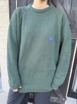 Buzo de punto verde oscuro, de manga larga con diseño raglán, cuello redondo acanalado y un pequeño parche bordado en el pecho.