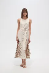 Vestido largo sin mangas, color crudo, con estampado de hojas en tonos marrones y negros. Presenta un cuello redondo y un lazo de tela a juego que ajusta la cintura. La falda es fluida y tiene aberturas laterales.