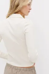 Top entallado corto de punto acanalado color blanco crema, con cuello camisero y escote en V. Tiene mangas largas.