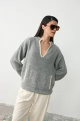 Sweater de tejido bouclé color gris, con cuello en V profundo y bolsillo frontal tipo canguro, ambos con detalles de borde en color crema. Presenta mangas largas y un corte holgado.