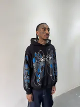Hoodie oversize de color negro con estampado artístico en pecho y mangas en tonos azul y gris. Presenta capucha forrada con tejido a cuadros, costuras reforzadas y grifa de la marca en la manga.