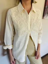 Camisa de manga larga en color beige, con cuello clásico y cierre frontal de botones. Presenta un delicado aplique de encaje tipo crunie en la parte superior delantera, aportando textura y un detalle artesanal al diseño.