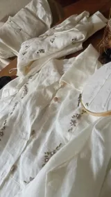 Camisa de lino color crudo con cuello clásico y cierre frontal de botones. Presenta bordados artesanales florales distribuidos por toda la prenda, realizados a mano.