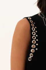 Chaleco negro de gamuza con diseño sin mangas, decorado con ojales metálicos plateados a lo largo de los bordes frontales y sisas.