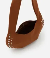 Cartera tipo baguette confeccionada en gamuza sintética color marrón. Presenta un diseño curvo con aplicaciones de tachas metálicas plateadas en los laterales, asa corta integrada y cierre mediante botón a presión.