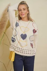 Buzo de punto color crema con estampado de corazones en tonos azul, rojo y amarillo, y detalles cuadrados en gris en el ruedo. Tiene cuello redondo y mangas largas.