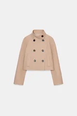 Chaqueta corta de paño en color beige, con cuello subido, manga larga y cierre frontal cruzado con botones oscuros. Presenta bolsillos delanteros de vivo.