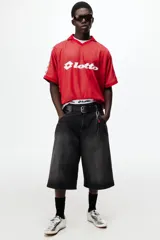 Camiseta de futbol de manga corta de la marca Lotto, en color rojo con un sutil estampado geométrico. Cuenta con cuello tipo polo abierto con escote en V y un calce holgado. Las mangas presentan una franja blanca lateral decorada con el logo de la marca repetido. El logotipo de Lotto destaca en blanco en el centro del pecho.