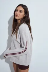 Sweater gris de calce relajado con cuello redondo y mangas largas. Presenta detalles de rayas horizontales en color rojo a lo largo de las mangas, con puños y ruedo acanalados.