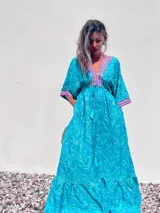 Túnica larga de estilo boho, color turquesa con estampado paisley en tonos azules y dorados. Presenta escote en V profundo con bordados decorativos en color fucsia y blanco, mangas amplias tipo kimono y ajuste en la cintura con cordón. El diseño incluye un ruedo con volado y terminaciones con borlas.