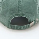 Gorra de tela verde con efecto desgastado, visera curva y parche rectangular frontal de cuerina marrón con diseño bordado de montaña y texto "Wild Life".