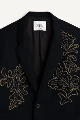 Blazer de corte relajado confeccionada en tejido con lana, color negro. Presenta cuello con solapas de muesca, cierre frontal de un botón y bolsillos de plastrón en la cadera. Destaca por su bordado en hilo dorado con motivos vegetales en la parte frontal.