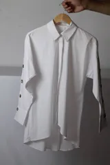 Camisa de lino y algodón color blanco, de corte oversize, con mangas largas abotonadas a lo largo del antebrazo y dobladillo asimétrico más largo en la parte trasera.