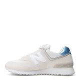 Championes urbanos New Balance modelo 574, color beige claro con detalles en blanco y azul metalizado en el talón. Confeccionados en una combinación de gamuza y malla, cuentan con el logo N característico en los laterales y entresuela con tecnología de amortiguación.