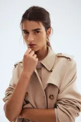 Gabardina de color beige claro, largo hasta la rodilla, de doble botonadura con botones marrones. Presenta solapas clásicas, hombreras, cinturón de tela extraíble con hebilla oscura y bolsillos insertados.