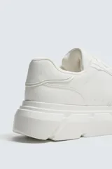 Zapatilla urbana de color blanco con diseño de suela gruesa con volumen y textura irregular. Presenta un corte superior con paneles combinados y cierre mediante cordones al tono.
