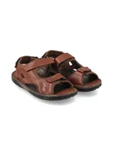 Sandalias de cuero color marrón, con diseño cerrado en el talón, tiras anchas en los laterales y cierre con velcro en el empeine y la parte frontal. Suela de goma negra con costuras visibles.