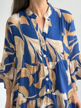 Vestido largo azul con estampado de hojas en tonos beige y blanco, escote en V con botones frontales, mangas largas amplias y diseño escalonado.