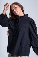 Camisa de color azul con cuello alto decorado con volados y lazo de ajuste. Presenta tablas decorativas en los hombros, cierre frontal con botones de nácar y mangas largas con puños elásticos que generan un efecto abullonado.