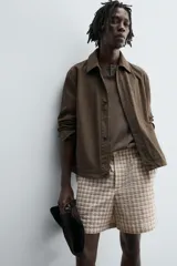 Bermuda de hombre, corte relajado, color beige con estampado de cuadros en tonos marrones y rosados. Presenta cintura elástica, bolsillos frontales y un bolsillo trasero de plastrón con cierre de botón.