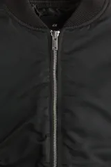 Campera bomber negra, ligeramente acolchada, confeccionada en nylon. Presenta cuello elevado acanalado, cierre frontal, bolsillos al bies con botón a presión oculto, bolsillo con cierre en una manga y dos bolsillos interiores con botón a presión. Los puños y el bajo son de remate acanalado ancho.