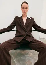 Blazer de corte sastrero en color marrón oscuro con diseño de rayas diplomáticas verticales. Presenta solapas clásicas, cierre frontal con botones y bolsillos laterales con solapa.