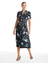 Vestido midi de seda fría con estampado floral en tonos azules sobre fondo negro. Presenta escote en V con cierre frontal de botones, mangas cortas con terminación fruncida, corte en la cintura con lazo de ajuste y falda plisada.