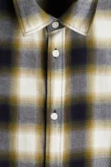 Camisa de franela de algodón suave con cuello inglés, cierre frontal de botones, mangas largas con puños abotonados y bajo redondeado. Presenta un diseño a cuadros en tonos oscuros y amarillentos.