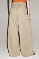 Pantalón de tiro medio confeccionado en hilatura de algodón, de corte wide leg y pernera ancha. Presenta cintura con trabillas, bolsillos laterales ocultos en costura, detalle de pliegues en la parte delantera y cierre frontal con cremallera, botón y ganchos metálicos.