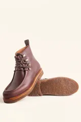 Bota de cuero vacuno color bordó con acabado charol, diseño de caña media con cordones y ganchos metálicos. Presenta costuras visibles en la puntera y suela de goma tipo crepe.