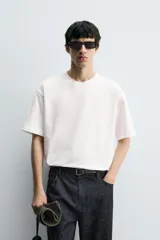 Camiseta blanca de corte holgado, cuello redondo y manga corta. Presenta un parche bordado en la espalda con diseño a contraste.