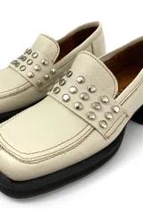 Mocasín color marfil de cuero, con detalle de tachas plateadas en la parte delantera y suela de goma negra.