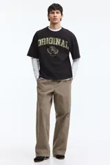 Remera negra de punto pesado de algodón con cuello redondo, hombros caídos y diseño holgado. Presenta un estampado frontal estilo universitario con la palabra "ORIGINAL" en tono desgastado y un gráfico circular rodeado de laureles.