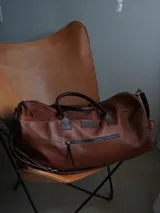 Bolso de viaje tipo weekender de cuero color tostado con asas y detalles en marrón oscuro, herrajes metálicos y bolsillo frontal con cierre.