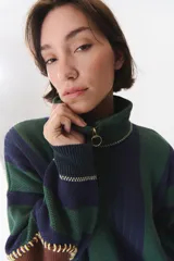 Buzo de punto con diseño de rayas verticales en tonos verde y azul marino. Presenta cuello alto con cierre frontal, hombros caídos y coderas en contraste de color marrón con costuras decorativas visibles.