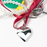 Llavero decorativo con mosquetón metálico en forma de corazón, que incluye una variedad de elementos colgantes: una pieza tejida a crochet con forma de sandía, una tira de perlas, cintas de raso rojas y cordones deportivos con punteras metálicas.