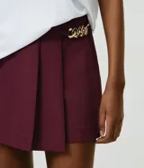 Short pollera de sastrería marca Cortelle, color bordó, confeccionado en crepé. Presenta sobreposición frontal efecto pollera con pliegues y detalle de cadena metálica decorativa en el lateral.
