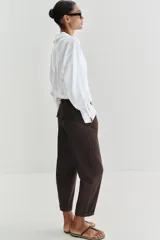 Pantalón chino de tiro bajo color marrón oscuro, con pernera recta y bajo con vuelta. Incluye un cinturón trenzado delgado de color marrón claro. Cuenta con bolsillos delanteros y cierre frontal con cremallera y botón.