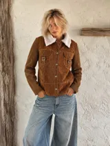 Campera de gamuza color marrón con cuello de corderito blanco, cierre frontal con botones metálicos y bolsillos frontales con hebillas decorativas.