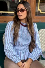 Blusa celeste a rayas marrones y blancas, con diseño de punto smock en el pecho y mangas largas abullonadas con puños elásticos.