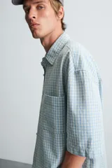 Camisa de corte relajado confeccionada en tejido de algodón, con estampado de cuadros pequeños en tonos celestes y blancos. Presenta cuello solapa, manga corta y bolsillo plastrón en el pecho con cierre frontal de botones.