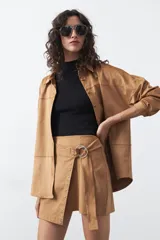 Minifalda de gamuza símil color tostado, con diseño envolvente, detalle de tachas metálicas en el lateral y cinturón integrado con aro metálico.