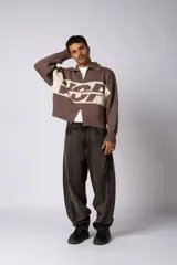 Campera tejida de punto con cierre frontal completo y cuello polo. Presenta un diseño de franja horizontal ancha en color crema con estampado gráfico oscuro sobre una base de color marrón. Posee puños y ruedo acanalados, con un pequeño parche de marca en el puño izquierdo.